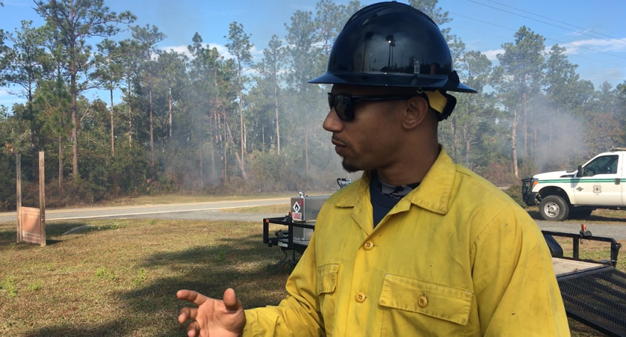 Man in black hat and yellow jacket looks behind him to a forest.