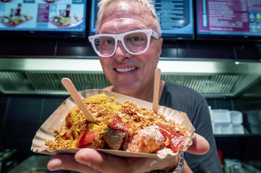 Lars Obendorfer, owner of "Best Worscht in Town" sausage, smiles holding a large sausage topped with curry powder and popsicle sticks standing up in it as utensils.