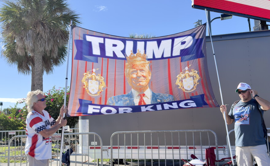 Some attendees waiting in line hold signs and flags, one of which reads "TRUMP FOR KING."