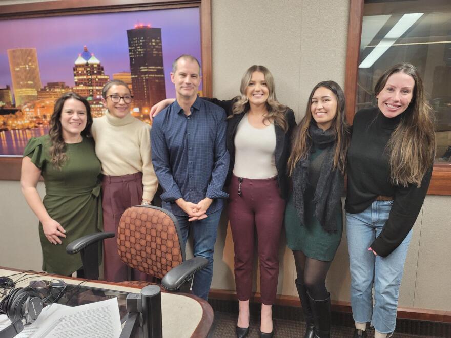 Six people stand with their arms around each other in front of a wall in a radio talk studio: a woman at far left has long brown hair and is wearing an olive green dress; a woman at near left has very short brown hair and is wearing glasses, a beige turtleneck sweater and pink pants; a man to her left has very short dark hair and is wearing a navy button-down shirt and jeans; a woman to his left has long blonde hair and is wearing a black blazer, white shirt and maroon pants; a woman at near right has long brown hair and is wearing a black scarf, a green dress and knee-high black boots; a woman at far right has long brown hair and is wearing a black turtleneck sweater and jeans.