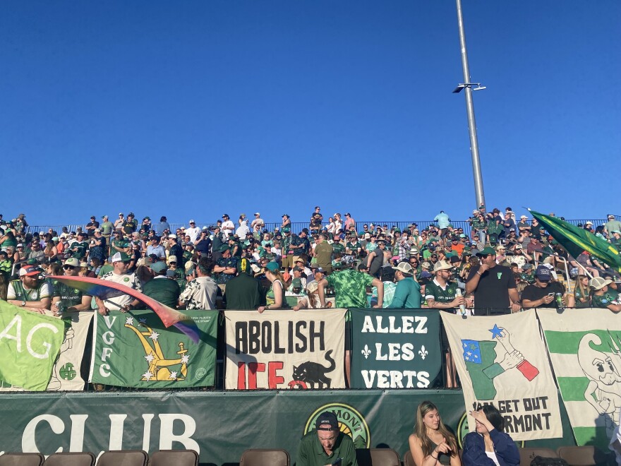 A crowd of soccer fans wave flags in front of banners in support of Vermont Green Football Club