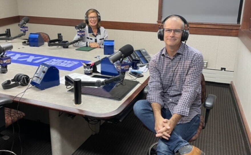 Two people wearing headphones and eyeglasses sit at a table in a radio talk studio: a woman at left has shoulder-length brown hair and is wearing a blue-and-white striped sweater with a blue collar; a balding man at right is wearing a plaid button-down shirt, blue jeans and brown shoes.