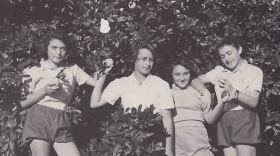 A black and white photograph of four young women posing together in an orchard in front of a leafy citrus tree laden with fruit. They are standing outdoors on small wooden boxes amid tall grass, wearing mid-20th-century casual shirts, shorts, and trousers while holding pieces of fruit.