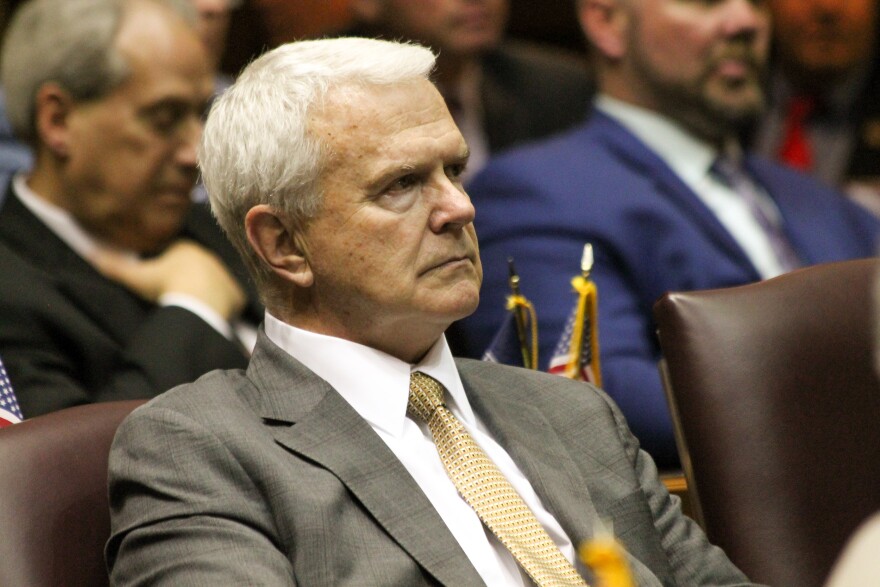 Indiana Representative Greg Steuerwald sits in a chair. He's wearing a suit and is looking off camera. Other men in suits sit behind him.