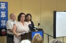 A woman in a white dress stands at a lectern facing out at a crowd of people seated at small tables. The backs of three women can be seen in the foreground. 