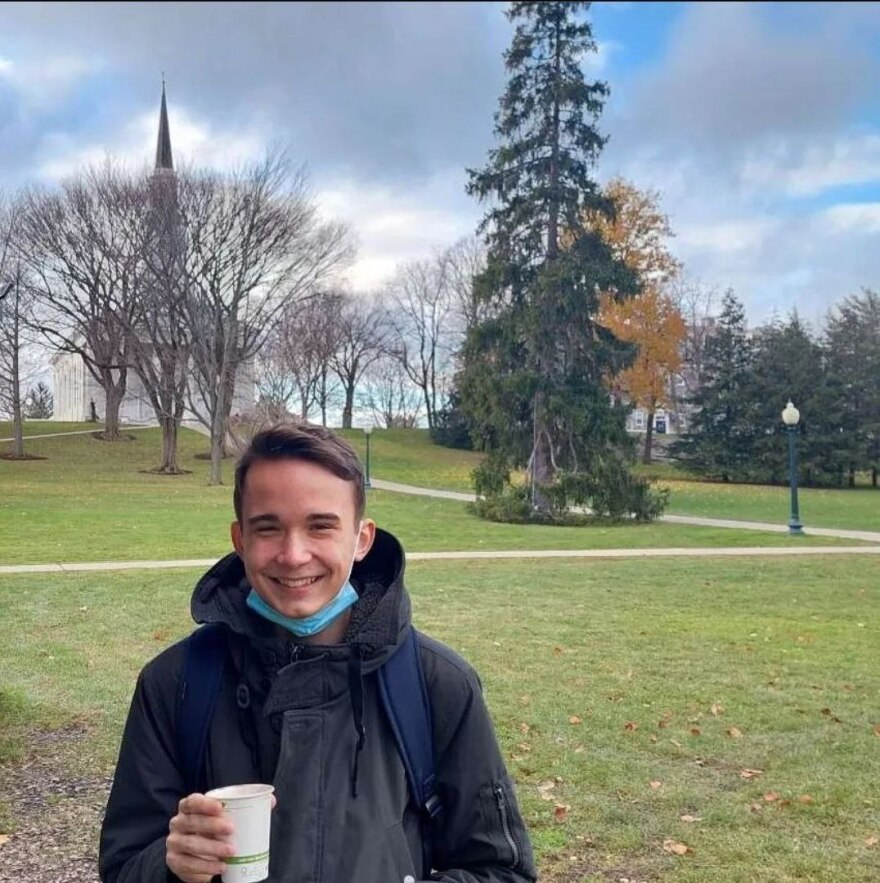 A young man in a black jacket holding a white coffee cup stands in front of Middlebury College's chapel.