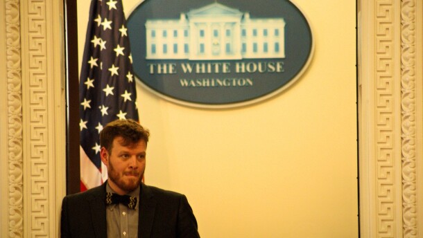 Zac Chase stands in front of an American flag and a sign that says "The White House."