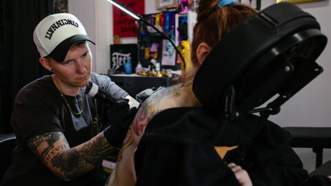 A person lays face down in a massage chair as another person tattoos their back