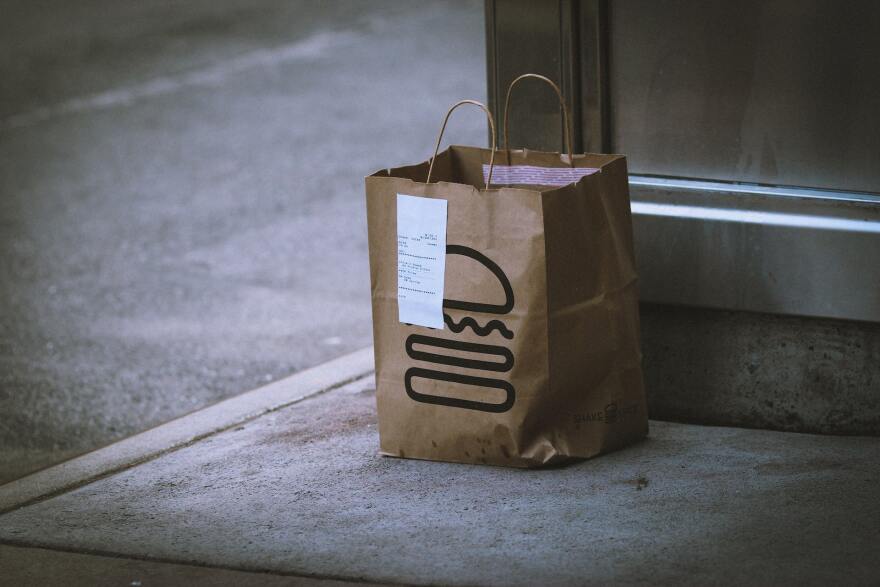 A brown paper bag decorated with a rudimentary illustration of a burger sits on a sidewalk corner, a receipt is attached