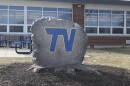 A large rock with the letters TV painted in blue with a gold outline placed next to a school building.