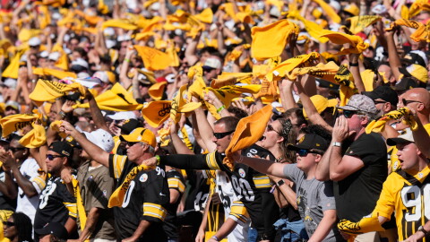 Pittsburgh Steelers fans cheer in black and gold standing in a filled section of a stadium waving terrible towels in a stadium 