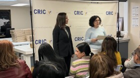 A small crowd sitting in white chairs and a woman in a black blazer watch a woman in a blue sweater speak.