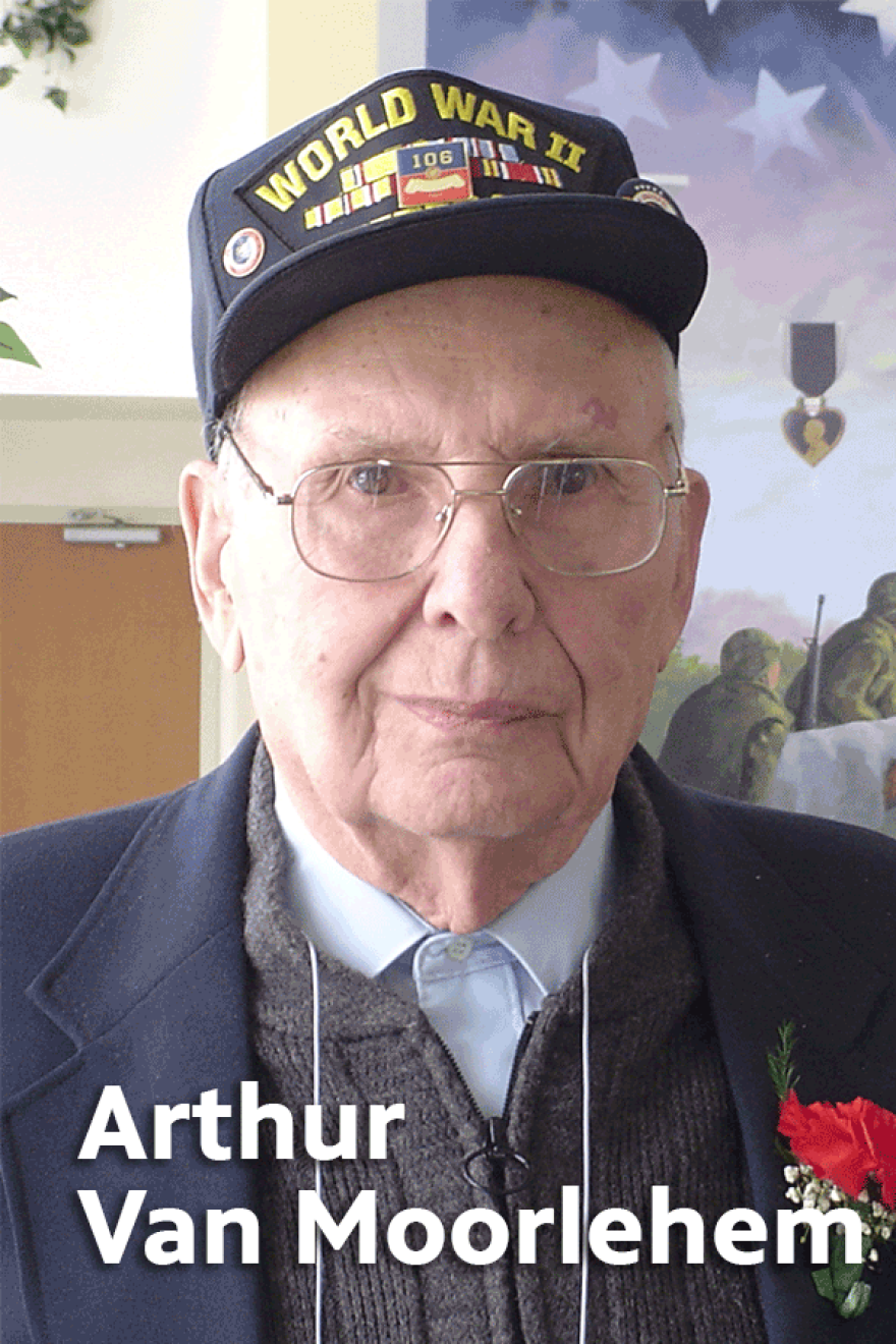 Elderly Arthur VanMoorlehem wearing glasses, a light blue shirt under a gray sweater, a navy blue jacket with a red carnation in the lapel, and a WWII baseball cap
