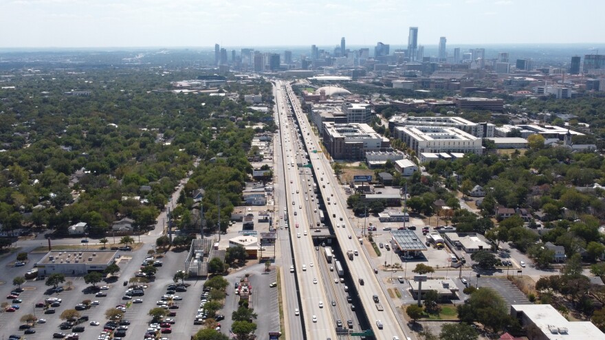 Vista aérea de la I-35 desde el Fiesta Mart. Las cubiertas superiores se extienden en la distancia. El horizonte de Austin se ve al fondo. Es un día claro y soleado, pero el horizonte está un poco borroso.