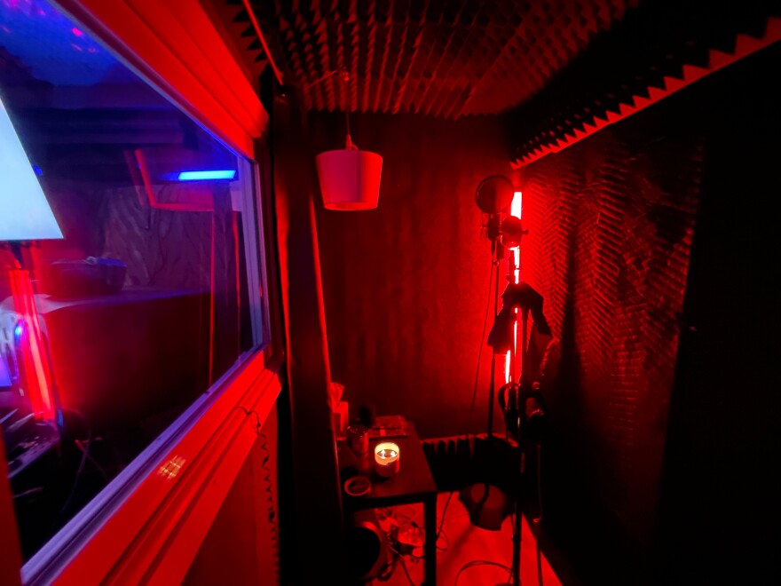 A dark recording booth with a red light in the corner and a microphone on a stand.