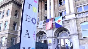 A white, vertical banner with the letters STEAM flies in front of the Syracuse City School District's newly opened STEAM High School's front steps.
