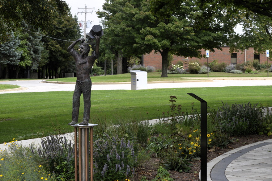 Bronze statue of a person holding a bucket overhead, standing on a pedestal in a landscaped garden area with trees, flowers, and a sidewalk nearby.