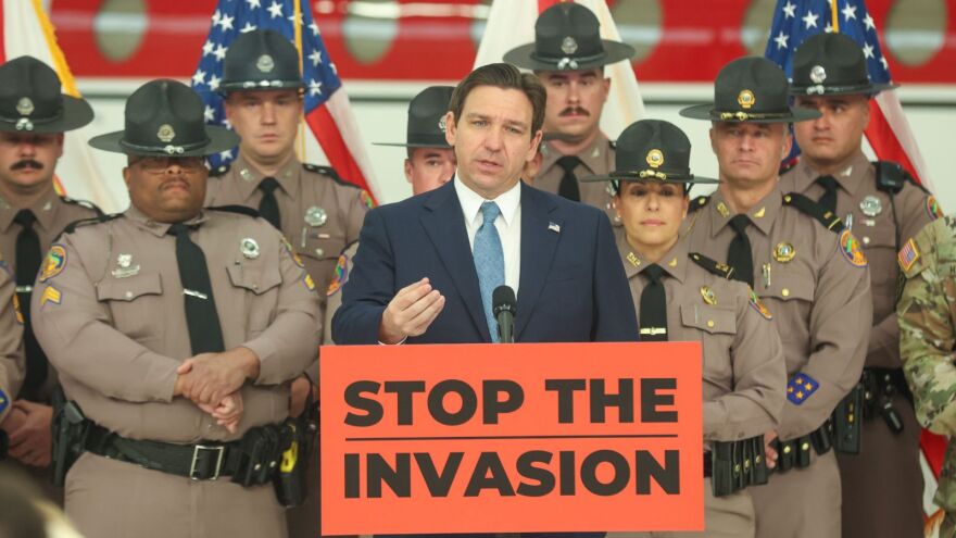 A man in a dark blue suit speaks at a podium, flanked by law enforcement officers in uniform