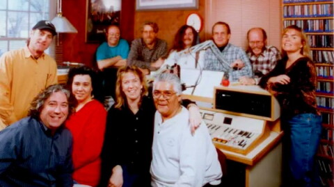 A group of 11 smiling people face the camera, they are seated and standing in a radio control room with a control board and a shelf of CDs in the back.