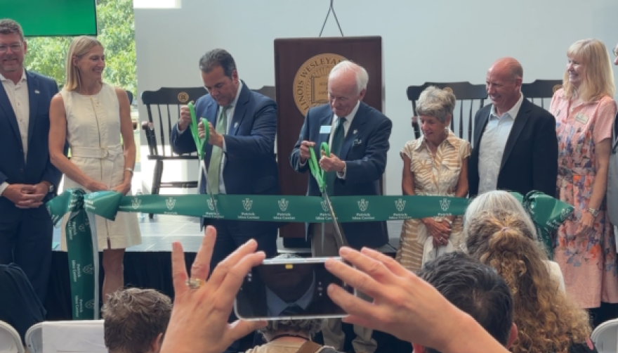 Two men in suits cut a green ribbon while several others look on