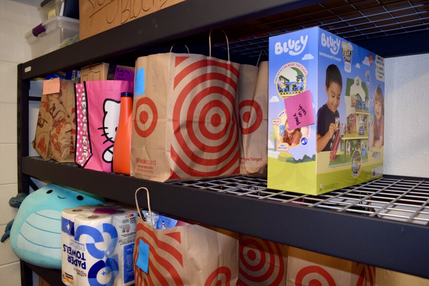 Shopping bags including one from Target and one with Hello Kitty are on a shelf with a Bluey toy.  The lower shelf holds a Squishmallow, a package of toilet paper and another Target bag. 