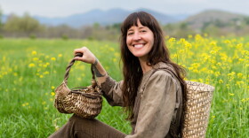 A lady sits in a field smiling with kudzu baskets and a white oak pack basket.