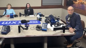 Three smiling people sit at a table in a radio talk studio: a woman at left has short blonde hair and is wearing glasses and a light blue top; a woman at center has long dark hair and is wearing a black top; a man at right has very short dark hair and is wearing glasses, a dark blue button-down shirt, jeans, and brown shoes