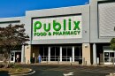 The front of a store says Publix Food & Pharmacy as seen from the parking lot. A woman in a blue nurse's uniform is walking to the left in front