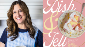 A woman in a blue apron smiles next to a copy of her new cookbook.
