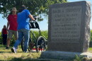 a memorial gravestone is in the foreground. In the background, a volunteering pushes an underground radar device while a man in a red t shirt watches