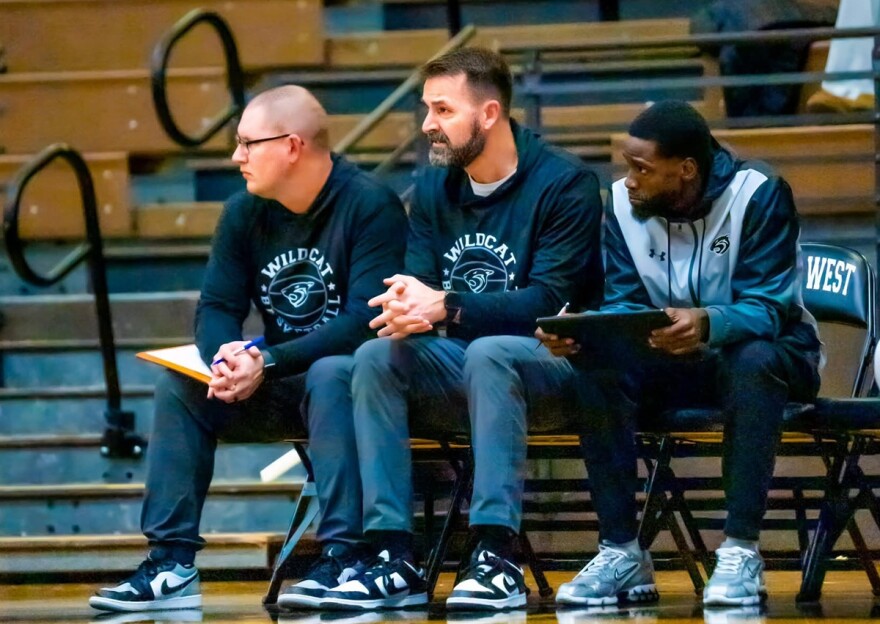 Three men seated next to each other in a basketball gymnasium