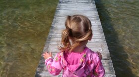 A little girl with blond ponytails and ap ink hooded coat walks on a long pier stretching into a body of water.