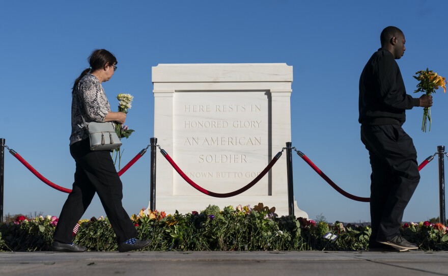 Two people holding flowers walk by the white marble Tomb of the Unknown Solider, against a blue sky.