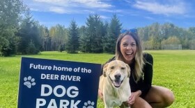 A woman poses with her dog on a green grassy field with a sign that says "Future home of the Deer River Dog Park."