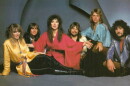 Six band members - two women and four men - sitting on the floor and looking into the camera. All have long hair
