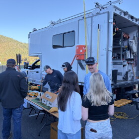 Students and presenters surround a tabletop model of power lines beside a utility truck at an outdoor career fair.