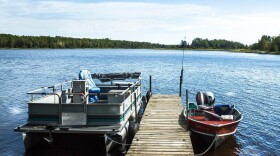  A pontoon boat and a red fishing boat are parked at a dock in a lake. A slight breeze ripples the water and green trees are visible in the background