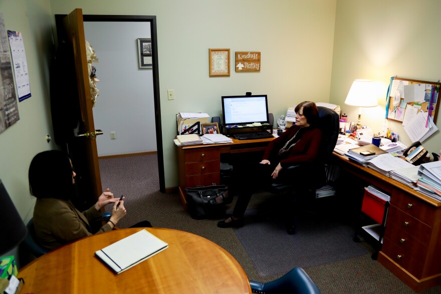 A woman sits in a swivel chair at a desk facing another woman sitting at a round table.