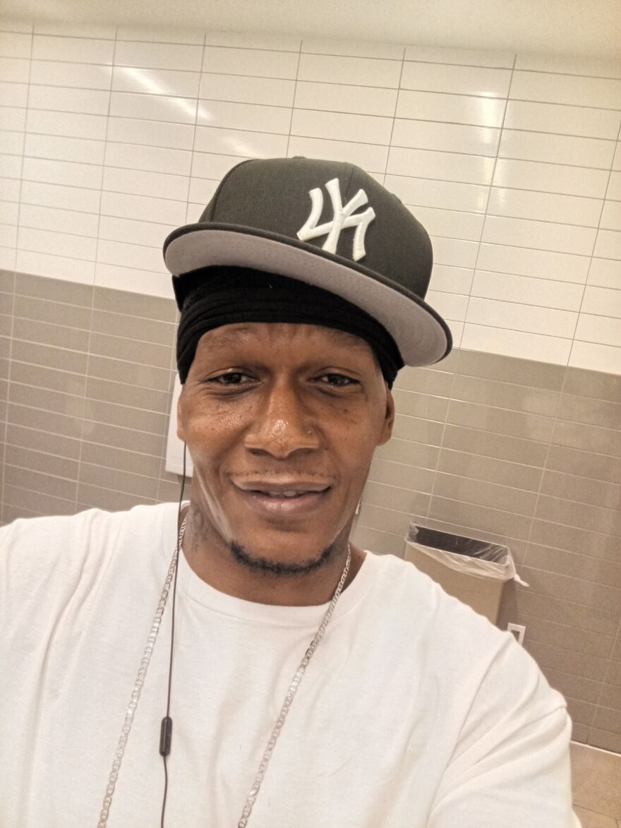 Man with New York Yankeets hat and gold chain, white shirt, standing against institutional gray tiled wall