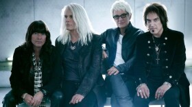 Four men with longer hair sit on a bench and look at the camera with neutral expressions on their faces.