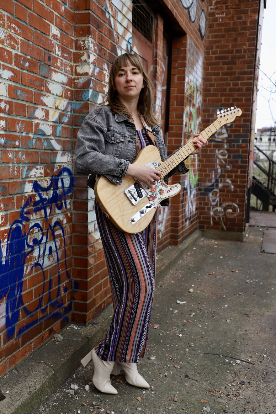 Bethany Joy stands against a brick wall holding a guitar