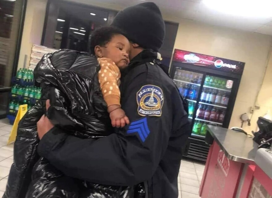 A police officer holds an infant boy with a coat wrapped around him in a pizza shop.