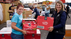 Two women, one with red hair in a bun on the left and First Lady Jennie Gordon on the right, smile while holding boxes of waffle fries. In the background, other people move boxes of food at a mobile food pantry.