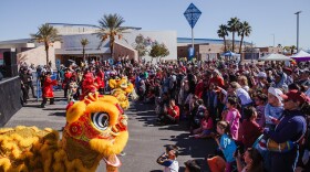A large crowd gathers outside the Desert Breeze Community Center on a sunny day to watch a traditional Chinese lion dance performance. Vibrant yellow and red lion dance costumes dominate the foreground, with performers in red traditional attire guiding the lions. Hundreds of spectators of all ages line both sides of the outdoor performance area, with children sitting cross-legged in front for a closer view. Palm trees, a "Desert Breeze Pool" sign, and event tents are visible in the background under a clear blue sky.