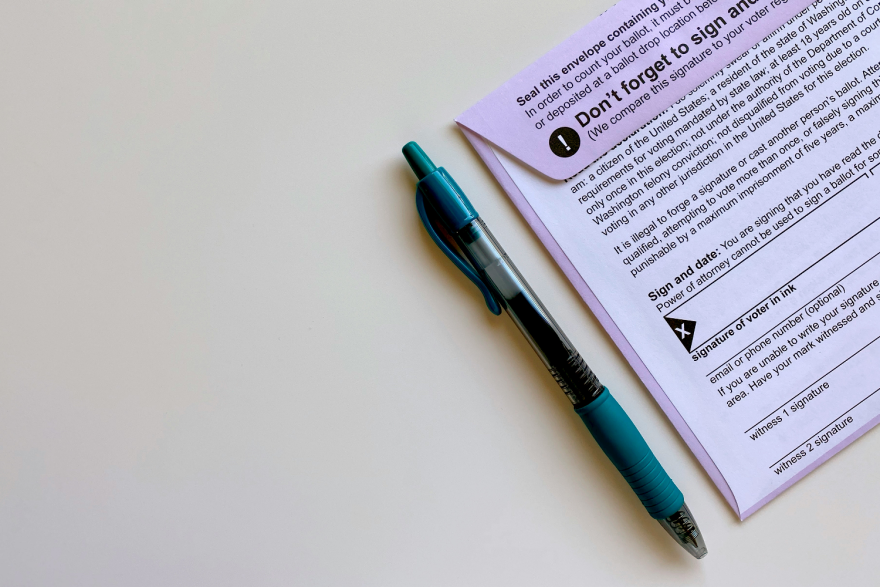 A mail-in ballot envelope with a pen sitting beside it. Some of the info on the ballot is visible, such as a reminder to sign the ballot and voting laws.