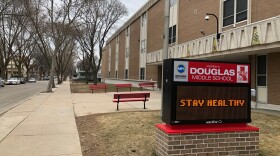 On the sidewalk outside a school building, an electric school sign says, "STAY HEALTHY" under the label "Andrew Douglas Middle School." 