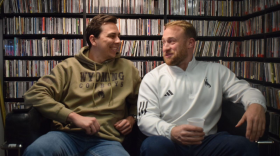 Two men sit on a bench together, looking at each other and smiling.