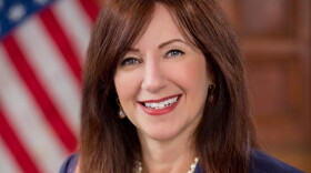 A woman with long brown hair, a blue blazer, and pearls smiling with an American flag in the background