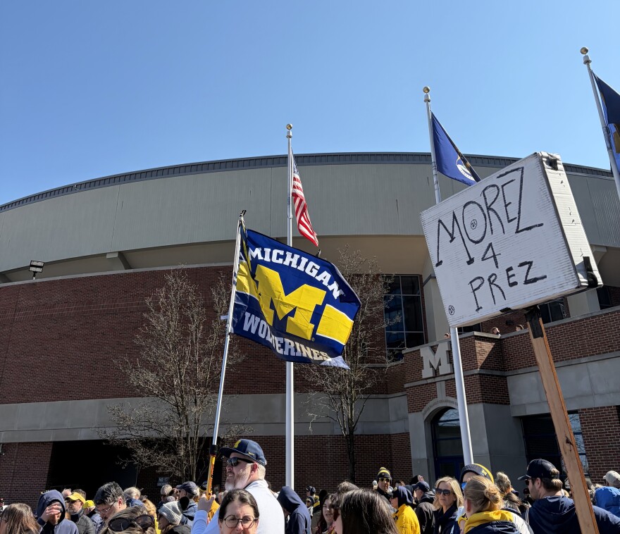 Michigan fans gather outside Chrysler Center to welcome home the 2026 National Champions. A flag supporting the Wolverines and a sign reading "Morez 4 Prez" is being held up.
