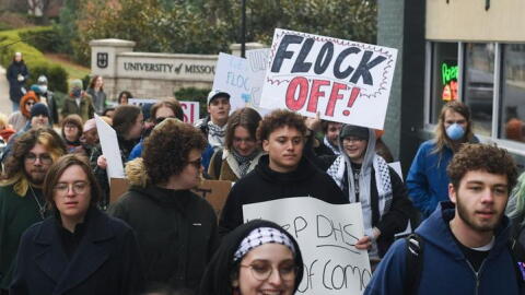 Mizzou students march down Ninth Street while carrying signs. One drawn in marker reads, "FLOCK OFF!" 